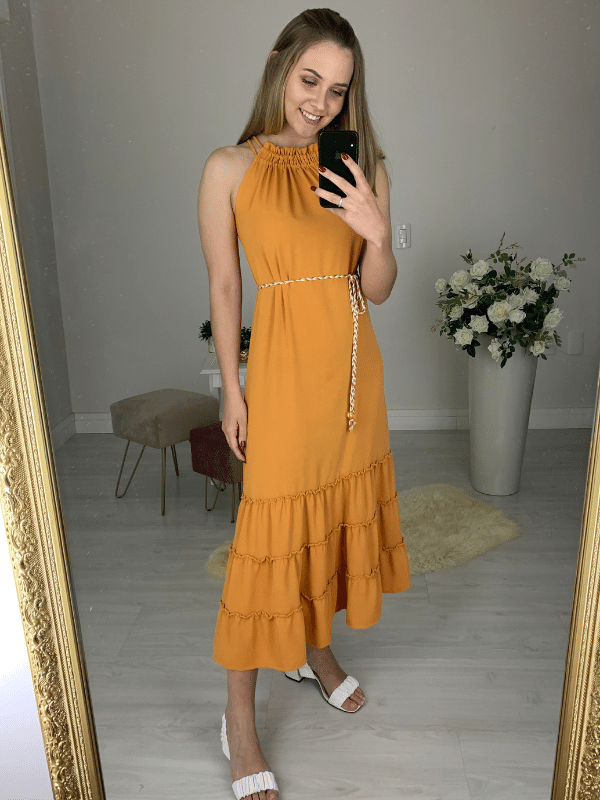 Vestido confeccionado em um tecido leve, fluido e que não amassa. Possui decote de amarração com detalhe franzido e dois cordões. Acompanha um cinto trançado das mesmas cores da peça. Sua saia tem comprimento midi e três babados. 